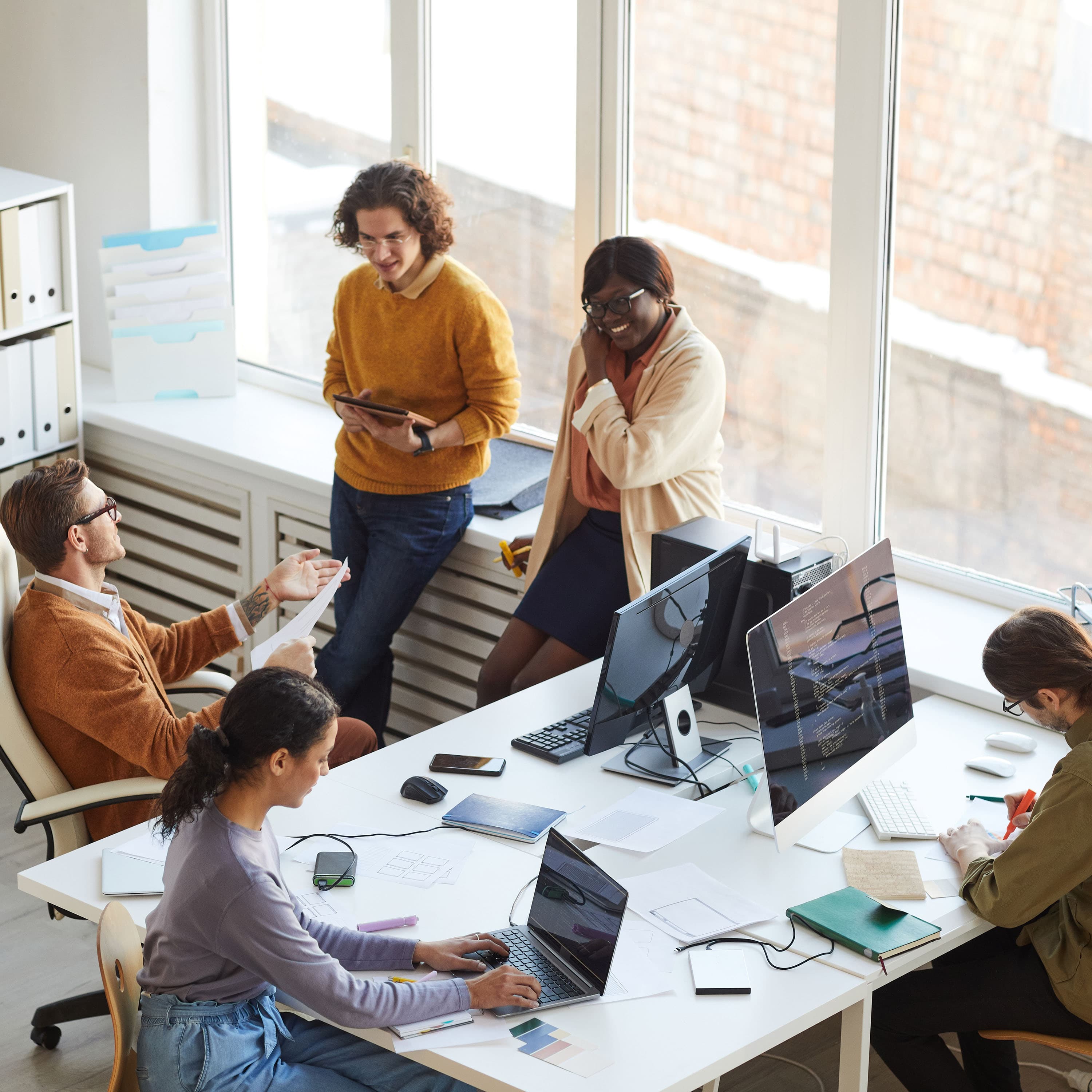 Ein vielfältiges Team von Fachleuten interagiert in einem hellen Büro; einige arbeiten an Computern, während andere im Gespräch sind.